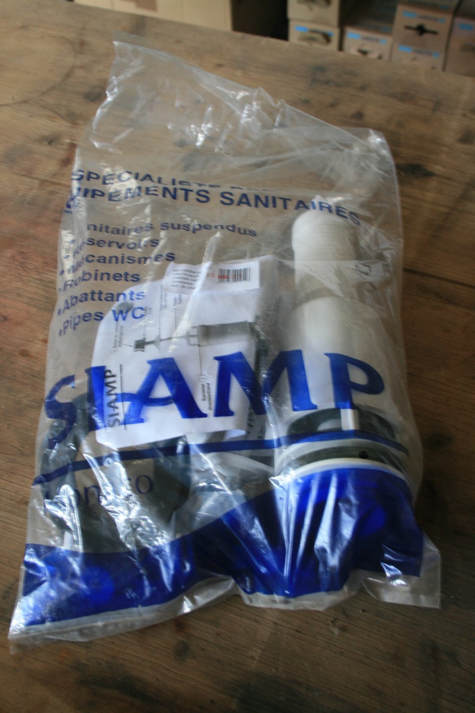 Sachet scellé SIAMP contenant un mécanisme de chasse d'eau à tirette chromé, posé sur un établi en bois