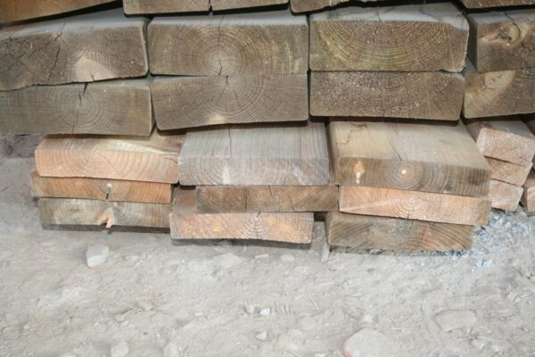 Pile de pièces de bois de charpente empilées, têtes de section visibles avec fentes de retrait et teintes grisées ou orangées