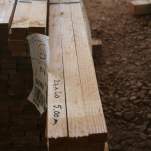 Pile de liteaux bruts de sciage posés sur du gravier, avec l'inscription au marqueur 27x40 5.00 m sur la pièce de droite