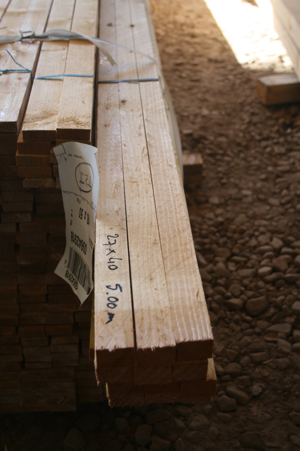Pile de liteaux bruts de sciage posés sur du gravier, avec l'inscription au marqueur 27x40 5.00 m sur la pièce de droite