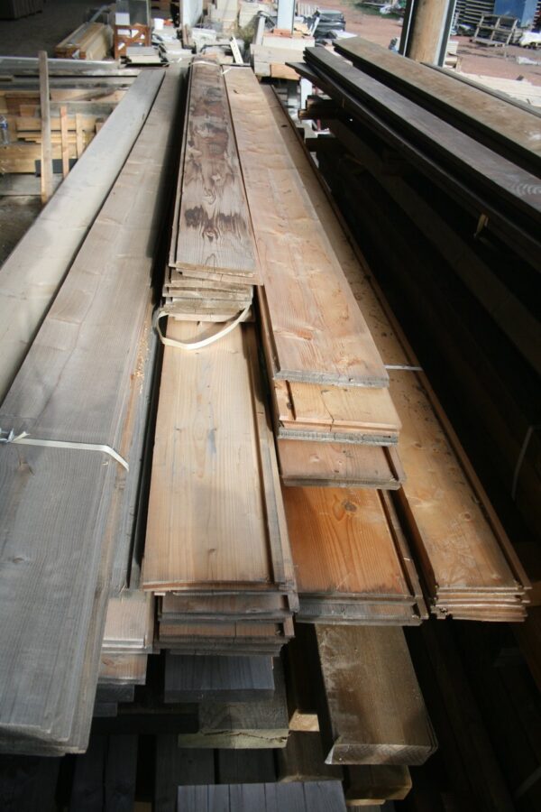 Piles de longues planches de rive en bois, brutes et de teintes variées, empilées sous un hangar à La Bricothèque